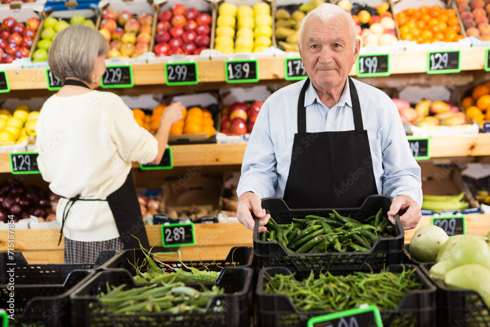 Elderly supermarket employee rearranges a box of green chili peppers