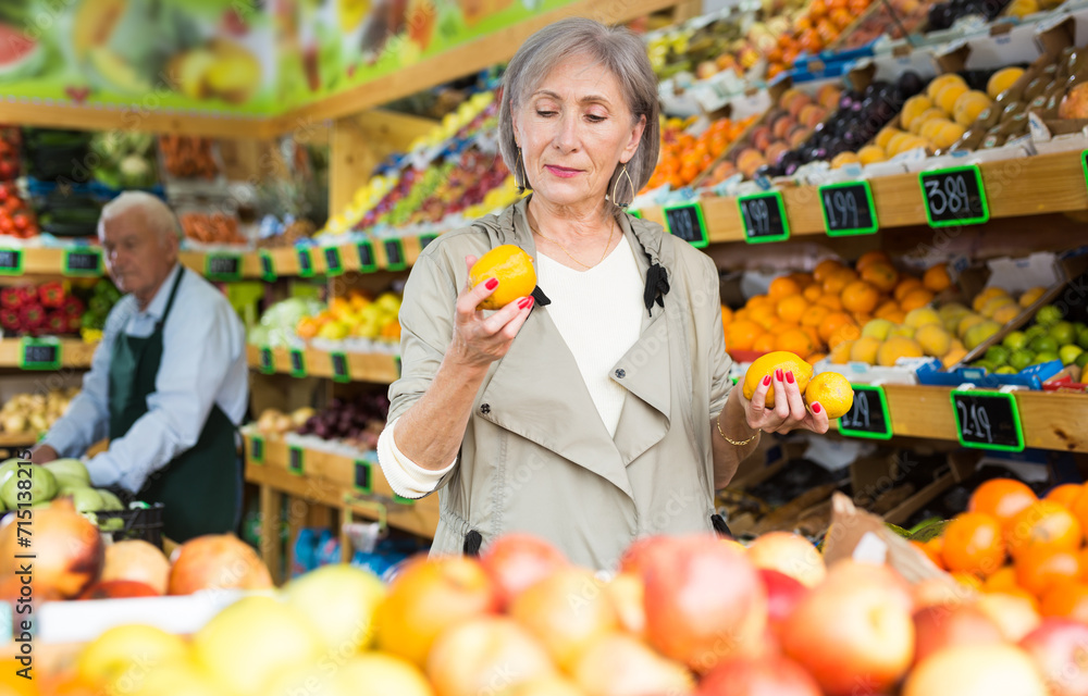 Old lady choosing fruits while standing in salesroom of greengrocer. Senior merhcandiser working in background.