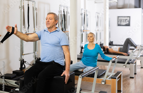 Serious old man in sportswear sitting and doing Pilates exercises for hands on reformer bed equipment in gym