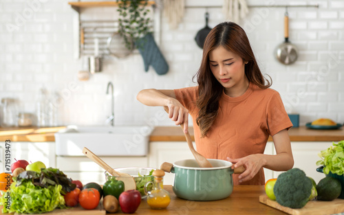 Young woman standing near stove and cooking, housewife, meal, chef, food.Happy woman looking and smelling tasting fresh delicious from soup in a pot with steam at white interior kitchen.