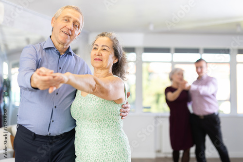 Positive aged woman and man practicing bachata dance moves in pair during group celebration in modern dancing studio