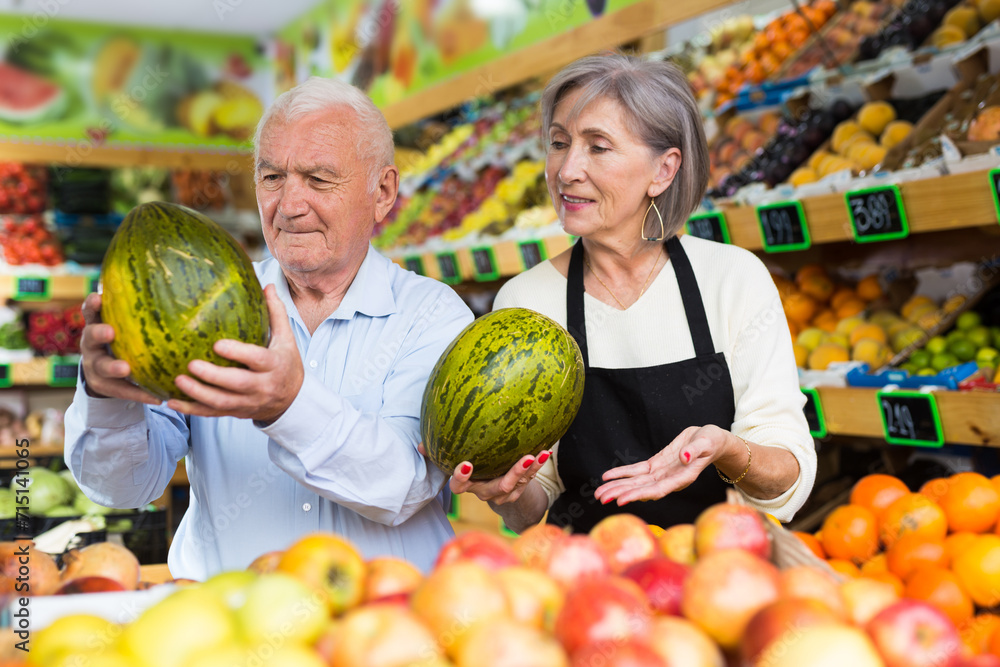 Fototapeta premium Woman supermarket employee helping mature man to choose watermelon and other fruits