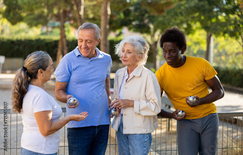 A group of mixed-race adult men and women of different age talking to each other and laughing during a break in petanque game in the city park on summer day