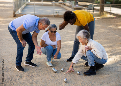 Group of multiracial mature people gathered together around silver metal boules/balls laying on the ground to measure distance between them while playing pentanque game outdoors in the park photo