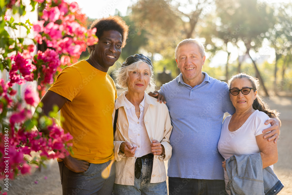 Cheerful age-diverse multiracial mixed-sex people in casual clothing posing outdoors in the park on a sunny day while standing together next to a flowering tree