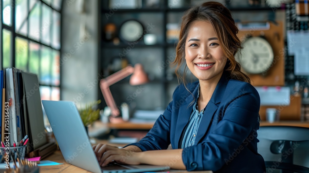 Smiling middle aged businesswoman in blue suit working on laptop at desk in office Smiling middle aged businesswoman in blue suit working on laptop at desk in office