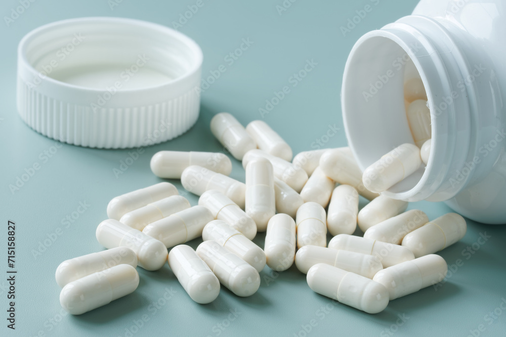White medical capsules next to open plastic container - bottle on pastel green-blue background. Photo. Selective focusing White medical capsules next to open plastic container - bottle on pastel green-blue background. Photo. Selective focusing