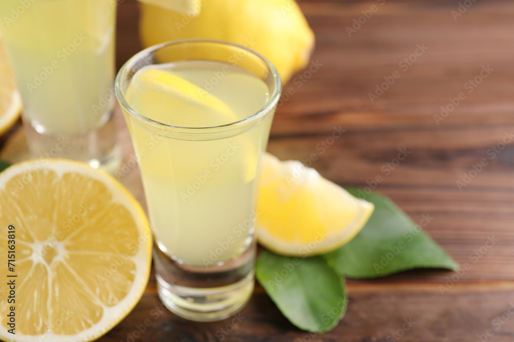 Tasty limoncello liqueur, lemons and green leaves on wooden table, closeup. Space for text