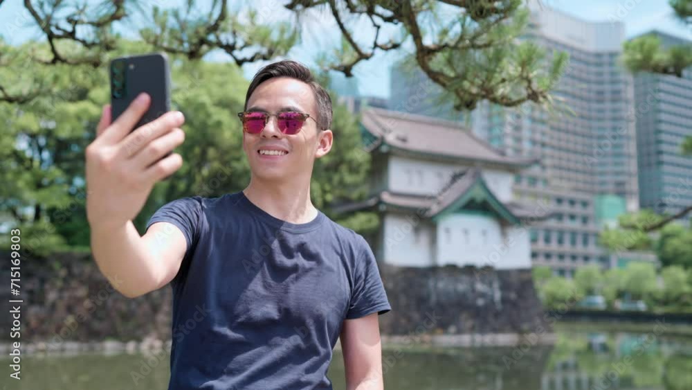 Positive young Hispanic male tourist with dark hair in casual clothes and sunglasses taking selfie on smartphone near of Imperial Palace located in Tokyo, Japan Positive young Hispanic male tourist with dark hair in casual clothes and sunglasses taking selfie on smartphone near of Imperial Palace located in Tokyo, Japan