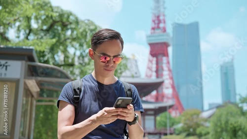 Smiling young Hispanic male traveler in casual wear and sunglasses with backpack smiling and messaging on smartphone while standing against ancient Zojo ji Buddhist temple near Tokyo tower, Japan photo