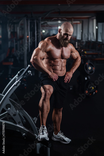 Muscular bald man posing in shorts. Bodybuilder showing off his shape in the gym. 