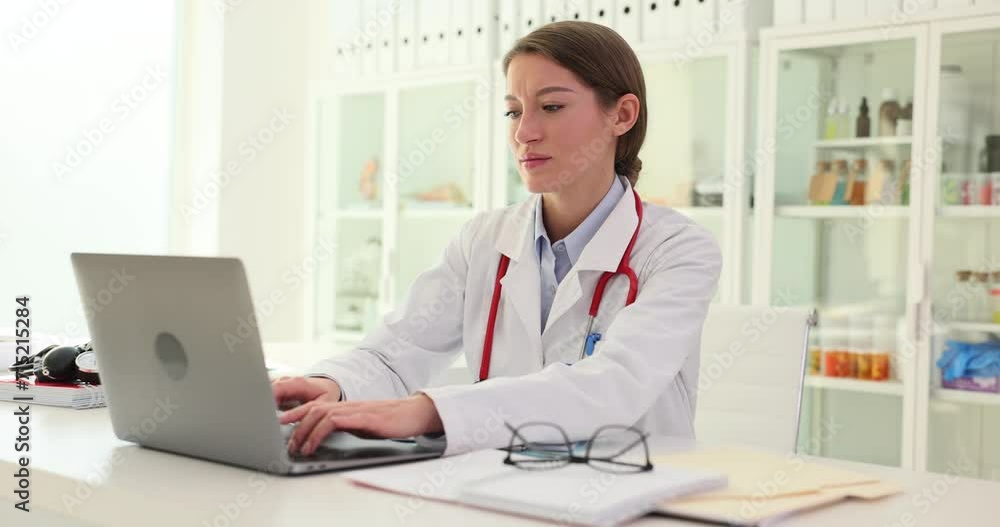 Attractive intern doctor works in hospital clinic office and uses laptop and completes paperwork Attractive intern doctor works in hospital clinic office and uses laptop and completes paperwork