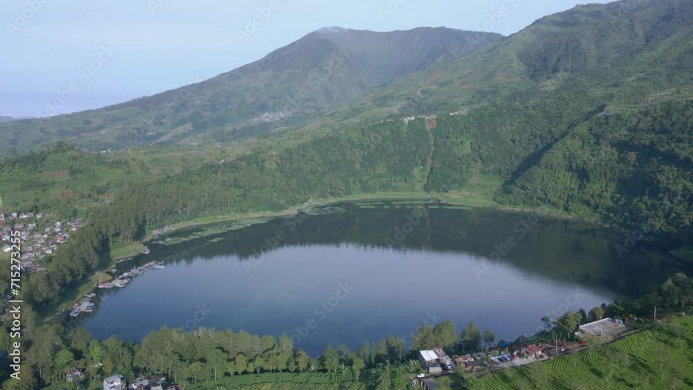 Aerial view of Menjer Lake. The crater lake with calm air is on a mountain slope with green tree vegetation. The surface of the lake reflects the sky and the shape of the hills and forests. Aerial view of Menjer Lake. The crater lake with calm air is on a mountain slope with green tree vegetation. The surface of the lake reflects the sky and the shape of the hills and forests.