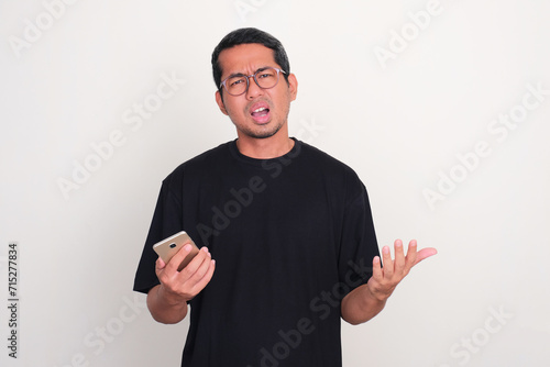 Adult Asian man showing disappointed expression while holding mobile phone photo