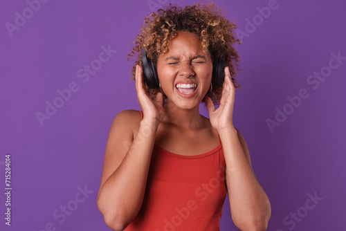 Young energetic cheerful African American woman in headphones squints eyes and sings loudly rejoicing at release of solo album of rock band member dressed in casual clothes stands on purple background