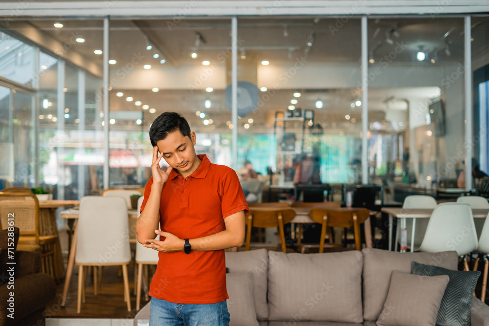 male shop assistant in red holding head while dizzy standing in furniture store male shop assistant in red holding head while dizzy standing in furniture store