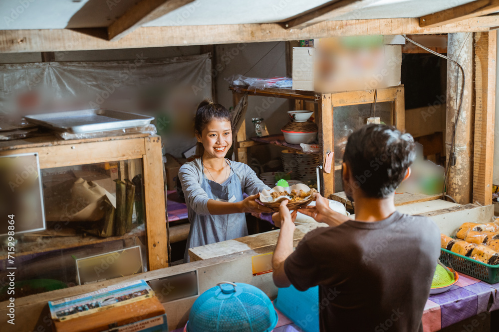 Asian woman selling a stall giving a plate of food to a buyer in the background of a stall