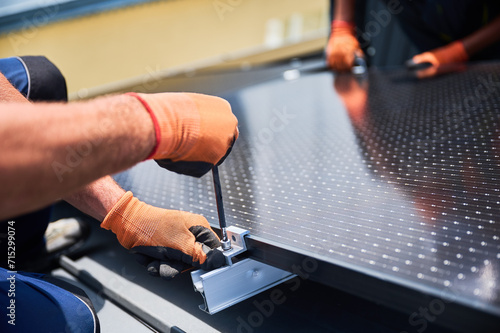 Worker building photovoltaic solar panel system on rooftop of house. Close up of man engineer in gloves installing solar module with help of hex key outdoors. Alternative energy generation concept.
