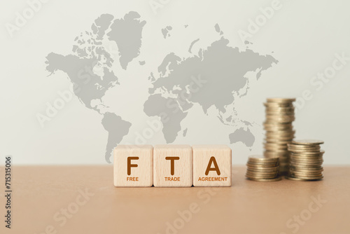 wooden cube blocks with the word FTA,  abbreviation for Free Trade Agreement, and blurred stack of coin on world map background