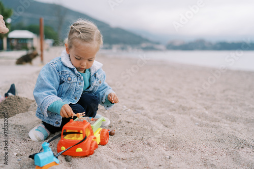 Little girl is squatting on the seashore and pouring sand into a toy truck