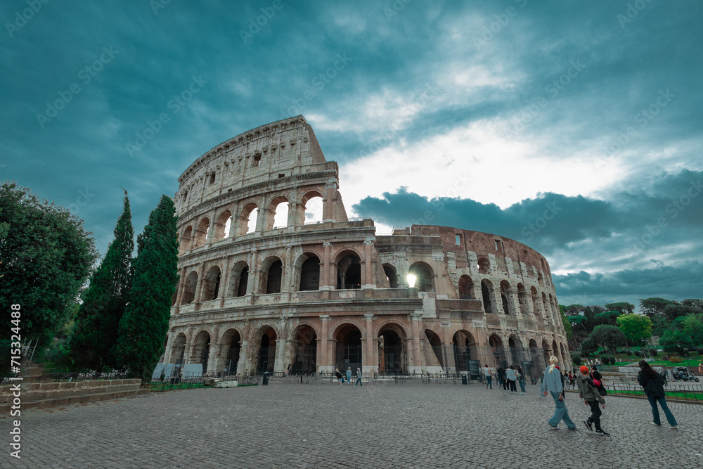 Early evening view of the colosseum in Rome, blue skies with dramatic clouds above the great famous amphitheatre. Autumn setting, some people around.