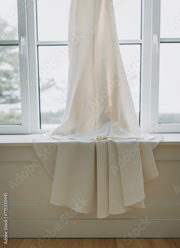 A white wedding dress hangs in a window and drapes over sill