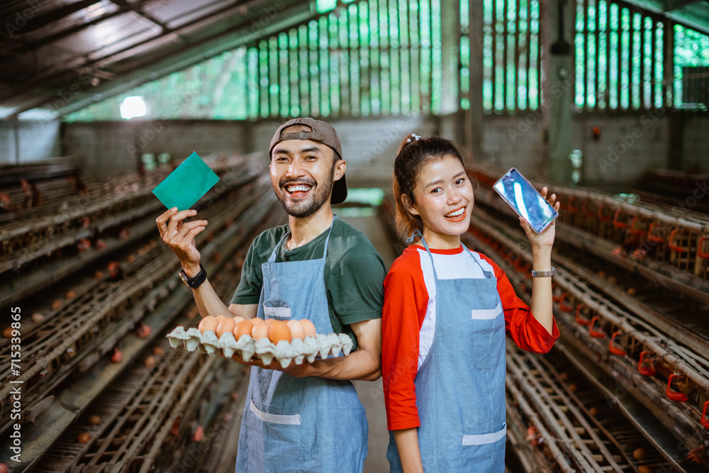 worker couple carrying cardboard tray for chicken eggs while showing passbook and cell phone stand at poultry farm
