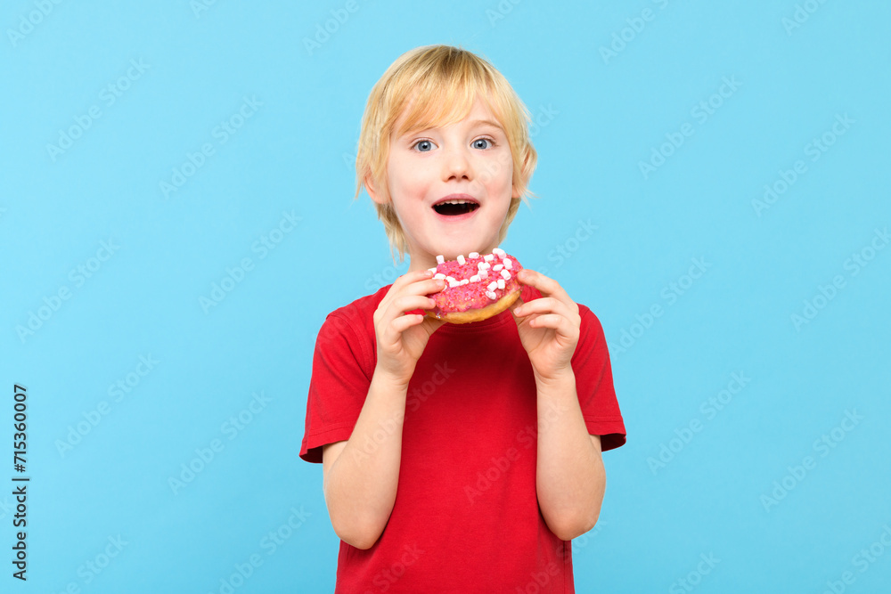 Cute little boy with blond hair and freckles eating a glazed donut. Children and sugary junk food concept. Boy holding colorful donuts, eating junk unhealthy food full of sugar.