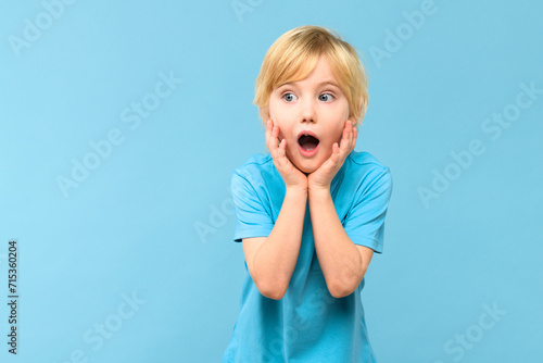 Wow! Portrait of a shocked cute little boy with blond hair on pastel blue background. Surprised preschooler studio shot, looking to side. photo