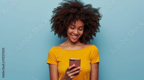 A young African American woman, radiating positivity, smiles while wearing a yellow top and using her smartphone against a light blue background photo
