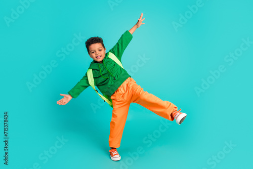 Full size photo of good mood small boy with rucksack wear green hoodie hold palms like wings isolated on turquoise color background