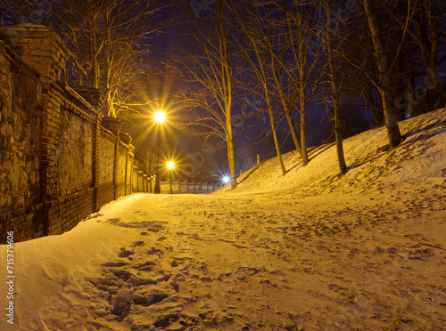 Park wieczorową porą, śnieg, latarnie oraz stary mur.