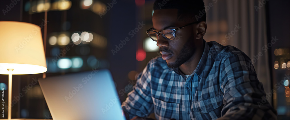 Man Working on Laptop Against Night Cityscape. Serious man in a shirt working late on his laptop with the glowing lights of the city skyline in the background. Man Working on Laptop Against Night Cityscape. Serious man in a shirt working late on his laptop with the glowing lights of the city skyline in the background.