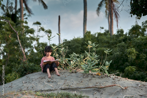 A cute little girl sits under a tree reading a book in the tropical forest