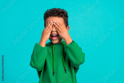 Photo of overjoyed schoolboy with wavy hair dressed green pullover arms cover eyes play peekaboo isolated on teal color background photo