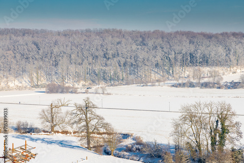 Winter landscape over the valley of Jekerdal just outside Maastricht, covered with fresh snow with a winding river through the scene and a forest in the distance on the slopes of a small hill 