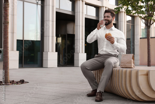 Businessman with hamburger and paper cup of coffee having lunch on bench outdoors