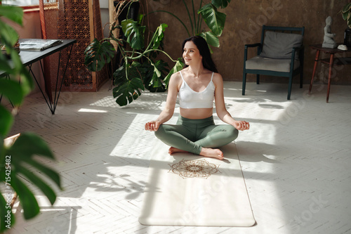 Content female practitioner peacefully performing padmasana in lotus position on cozy yoga rug. Charming woman sitting on floor of stylish exotic room and lloking to the window photo