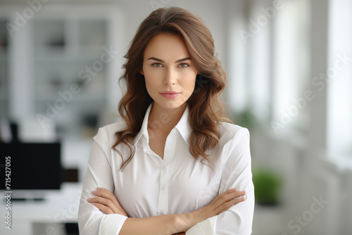Portrait photo of a beautiful businesswoman wearing a white shirt, folding her arms across her chest and looking forward at the camera. office background bokeh