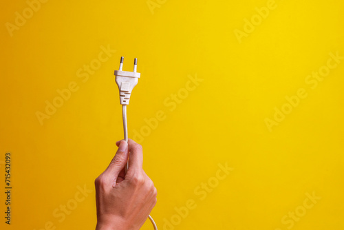 Hand holding white electrical plug isolated on yellow background with copy space. Save energy and earth hour concept photo