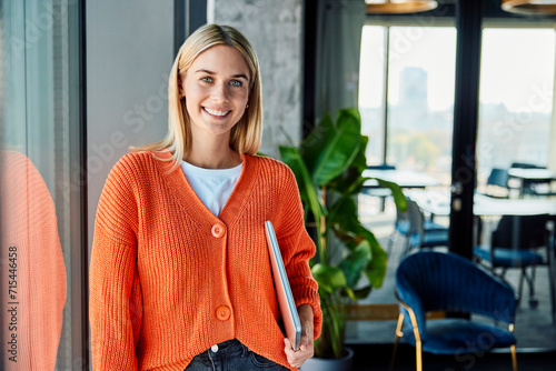 Happy young businesswoman standing with laptop near window in office photo