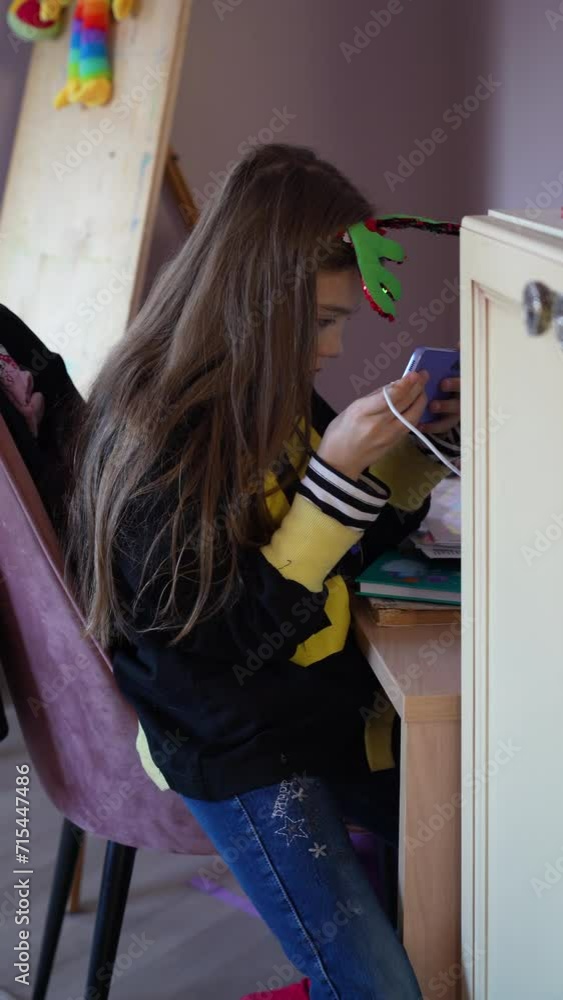 Teenage girl with long hair and reindeer antler headband is looking at her cell phone. She is wearing yellow and black sweater and has white charging cord attached to her phone. Teenage girl with long hair and reindeer antler headband is looking at her cell phone. She is wearing yellow and black sweater and has white charging cord attached to her phone.