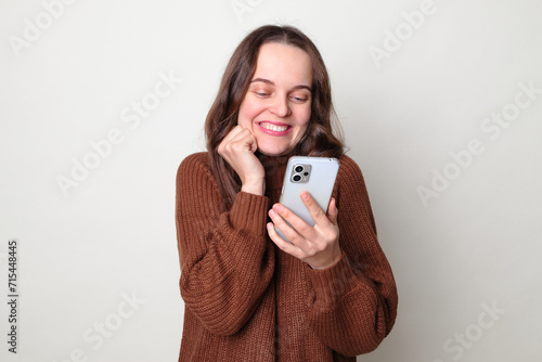 Pleased smiling delighted satisfied Caucasian woman wearing brown jumper standing isolated over gray background using smartphone reading pleasant message chatting online