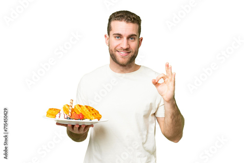 Young handsome blonde man holding waffles over isolated chroma key background showing ok sign with fingers