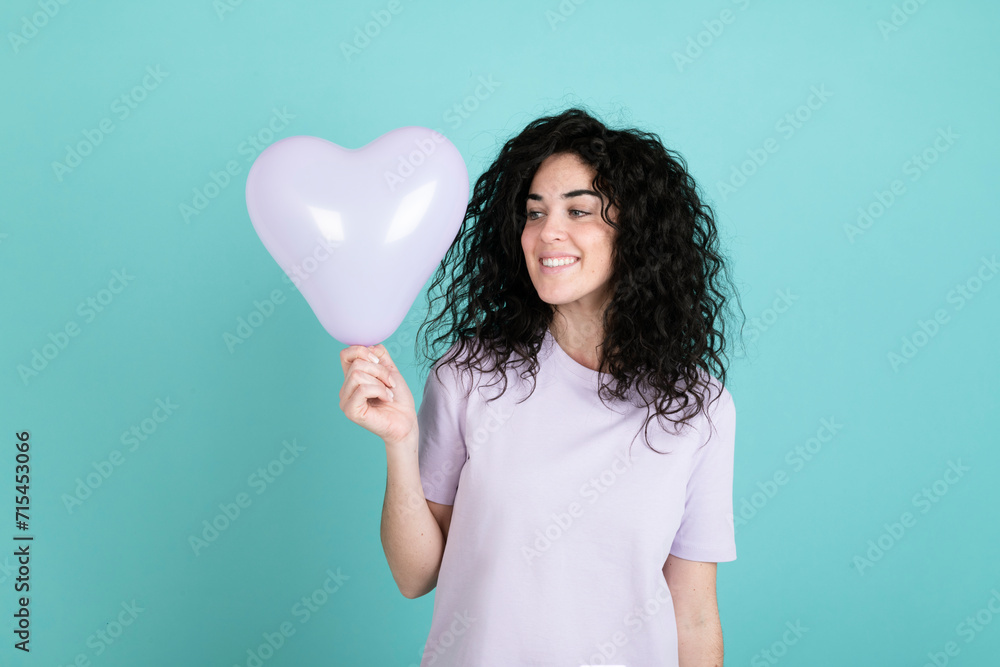Smiling woman holding pink heart shaped balloon against turquoise background Smiling woman holding pink heart shaped balloon against turquoise background