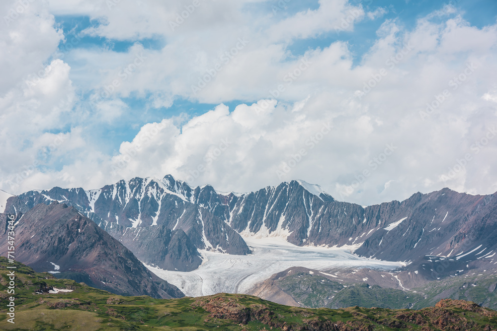 Dramatic alpine layered landscape with green hills and rocks with view to big glacier and large snow-capped mountain range far away under clouds in blue sky. High snowy mountains in changeable weather Dramatic alpine layered landscape with green hills and rocks with view to big glacier and large snow-capped mountain range far away under clouds in blue sky. High snowy mountains in changeable weather