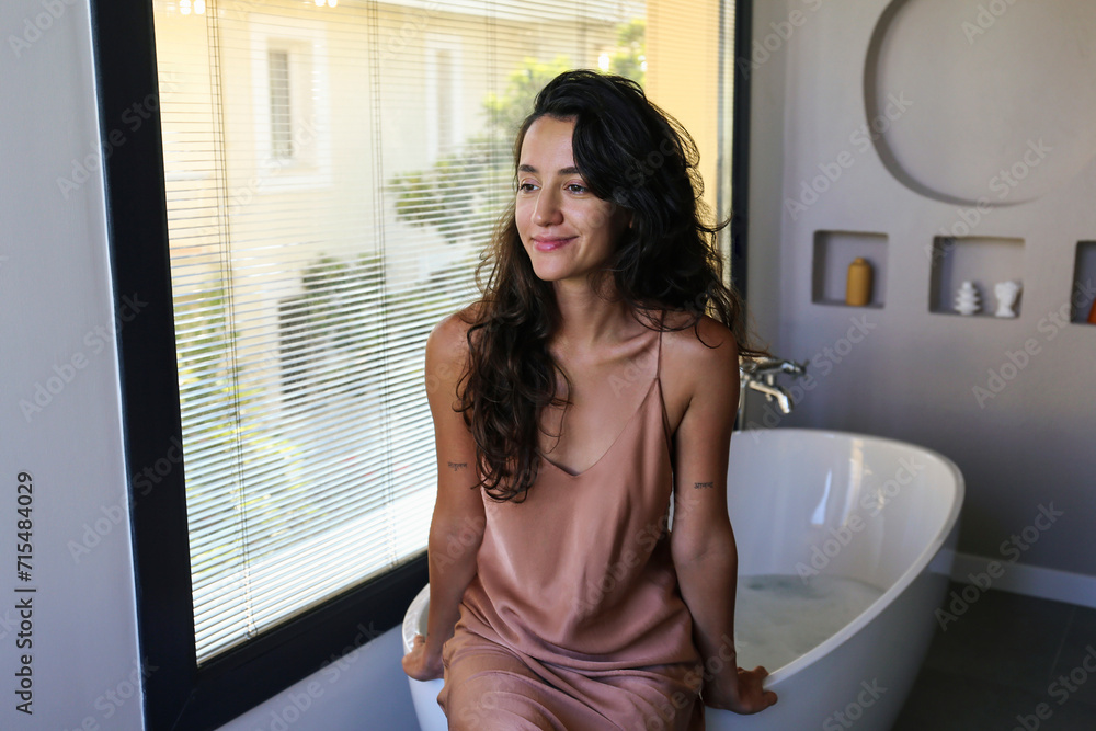 Young beautiful woman with brunette hair about to take a bath. Brunette female wearing silk nightgown sitting on the edge of the bathtub. Close up, copy space, background. Young beautiful woman with brunette hair about to take a bath. Brunette female wearing silk nightgown sitting on the edge of the bathtub. Close up, copy space, background.