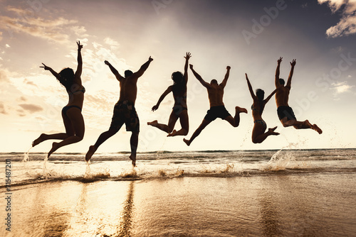 Group of silhouettes of happy young friends are having fun and jumps together at sunset sea beach