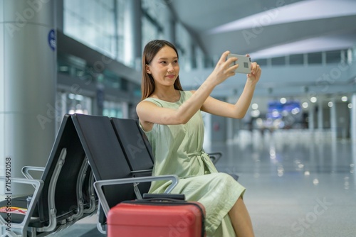 Asian woman with luggage using phone taking a selfie while waiting for departure at a transportation departure hall