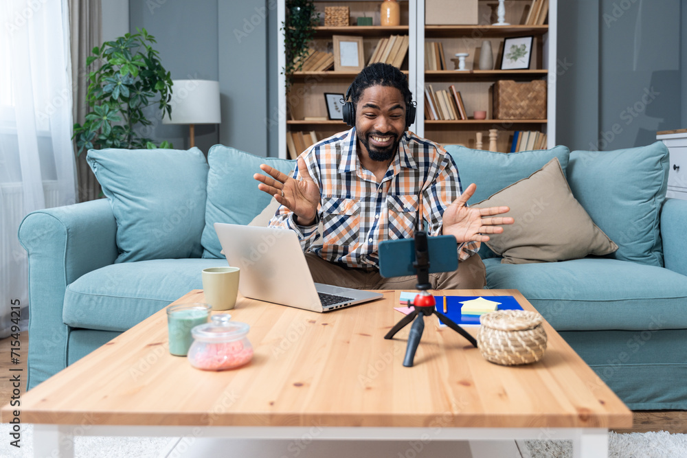 Young African American man with headset having web call using laptop computer and smartphone sitting on sofa talking at webcam in home office Video conference online business product strategy meeting Young African American man with headset having web call using laptop computer and smartphone sitting on sofa talking at webcam in home office Video conference online business product strategy meeting